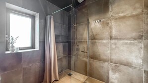 Una ducha con puerta de vidrio, una ventana con cortina y suelo de baldosas.