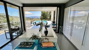 A modern dining area with a glass table, chairs, and a view of a pool and landscape. - MAGNIFICENT VILLA WITH SWIMMING POOL AND SEA VIEW FOR 8 GUESTS (GRIMAUD)