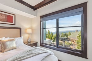A hotel room with a large window, a bed with white linens, a nightstand with a lamp, and a framed picture on the wall. - Constellation Luxury Residences (Truckee)