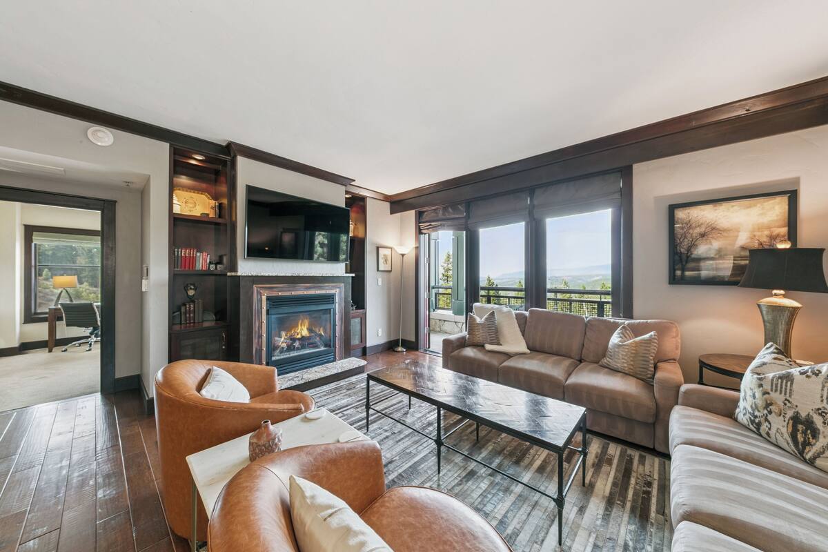 A spacious living room with a fireplace, leather sofas, a coffee table, and large windows offering a view.