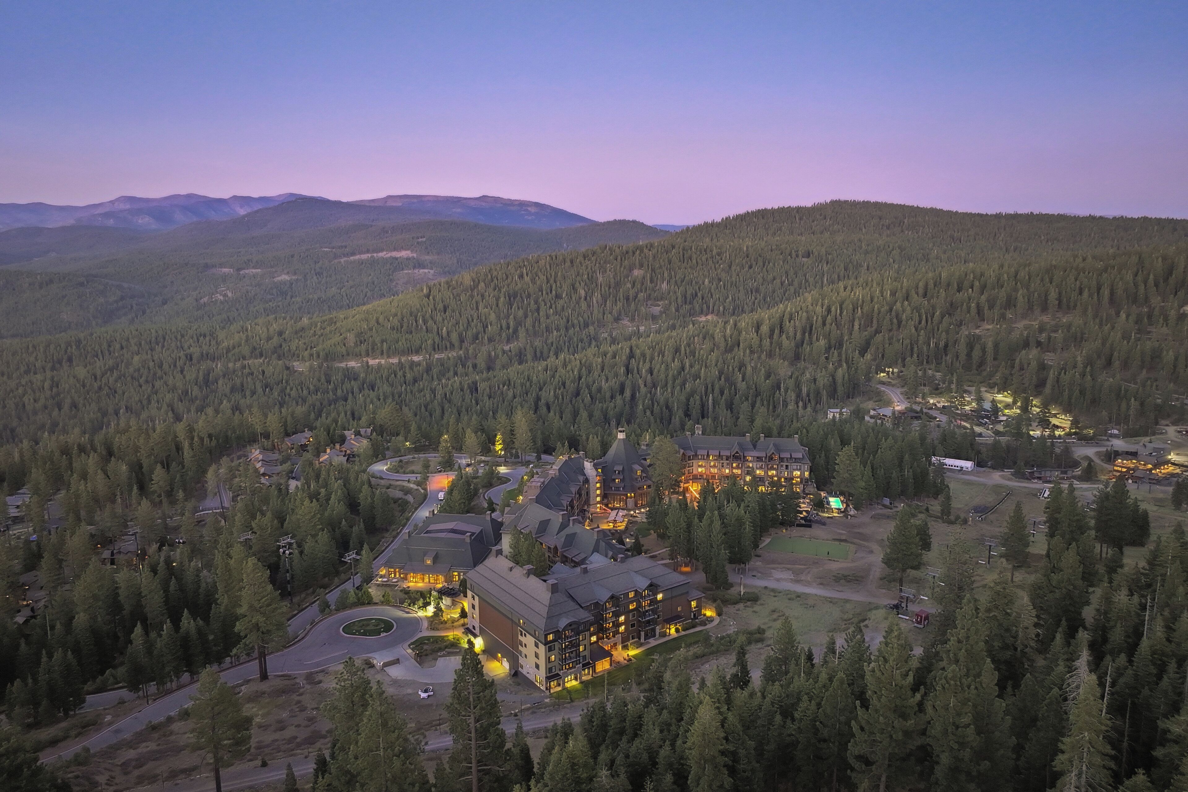 Vue aérienne d’un chalet composé de plusieurs bâtiments, entouré d’une forêt dense et avec des montagnes en arrière-plan.