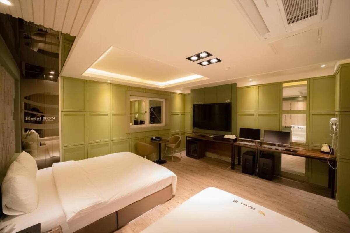 A hotel room with a bed, a desk with a TV, a mirror, and a kitchenette.