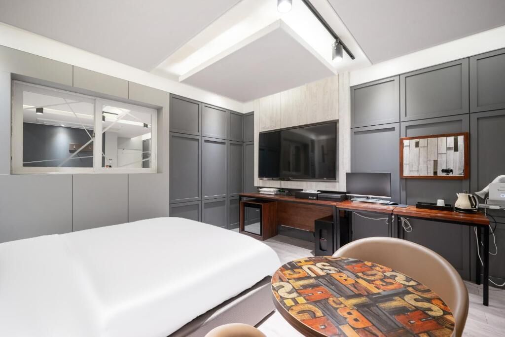 A modern hotel room with a bed, a desk with a flat-screen TV, a small table, and a chair.