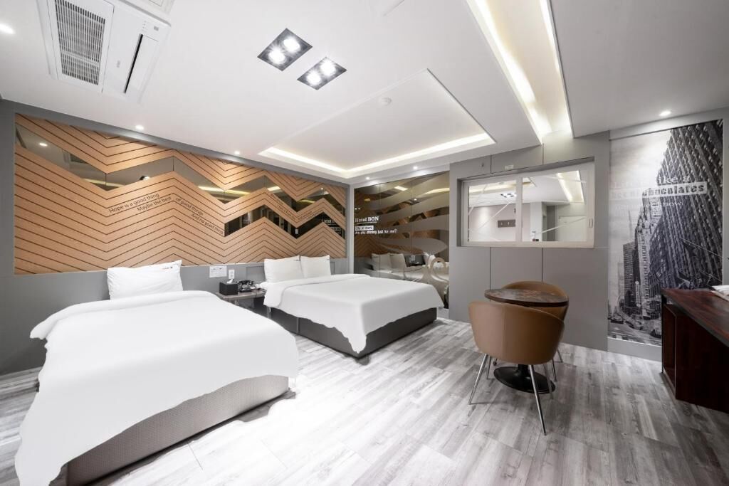 A modern hotel room with two beds, a seating area, a mirror, and a cityscape mural.