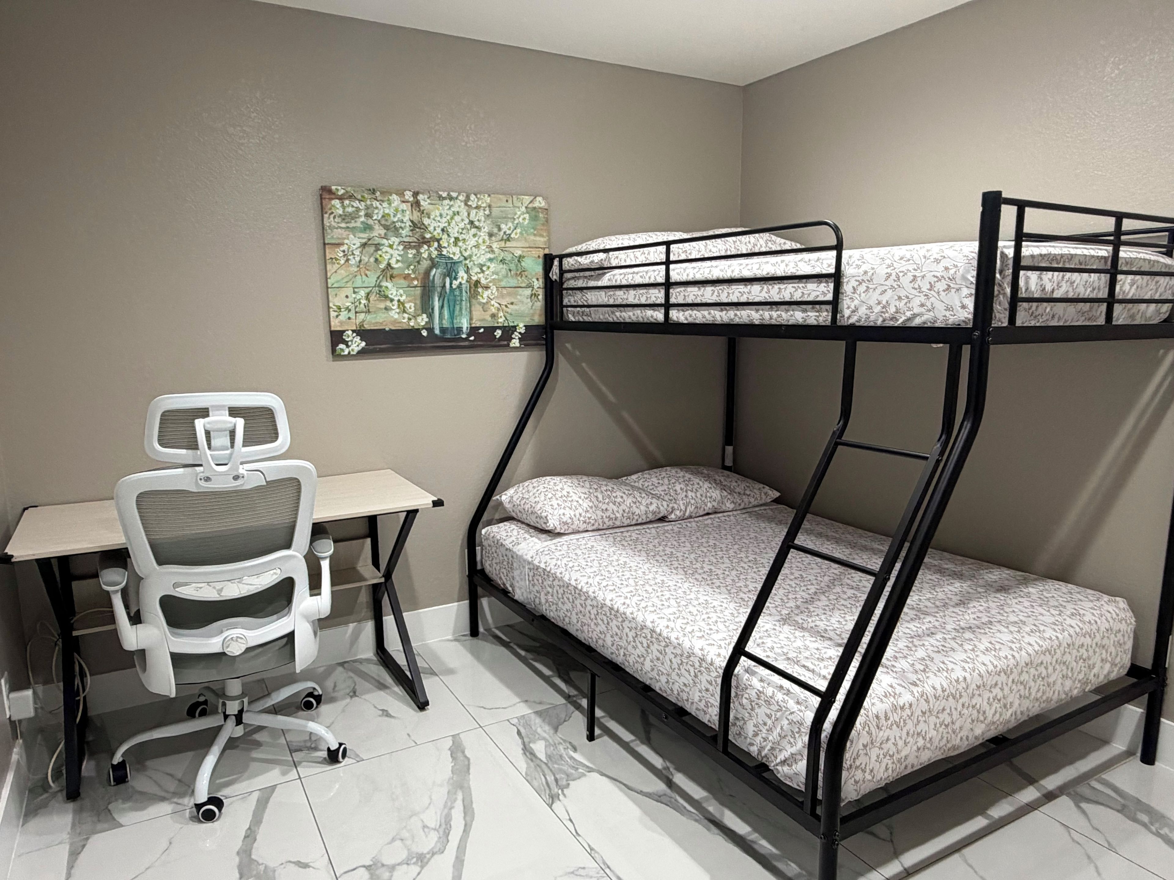 Un lit superposé avec un bureau et une chaise, un tableau accroché au mur et un sol carrelé.