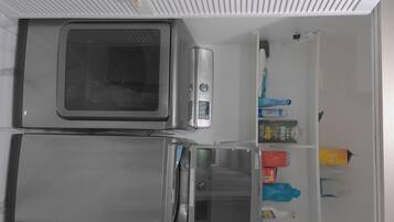 Una cocina con microondas, refrigerador y una estantería con diversos objetos, incluyendo una botella y una caja.