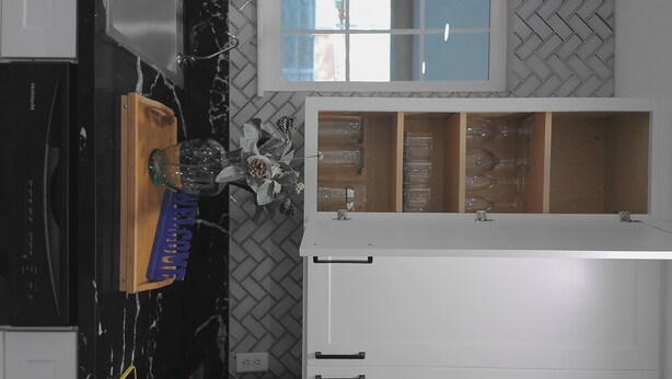 Cozinha com backsplash em padrão herringbone, janela sobre a pia e armário de madeira com a placa 'WELCOME'.