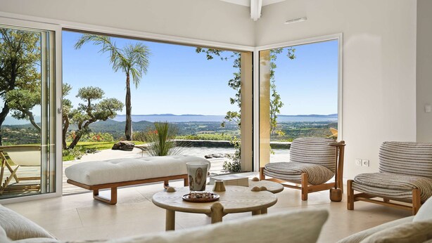 A modern living room with a large glass door leading to a scenic outdoor view, a cushioned bench, a round table, and striped armchairs. - Air-conditioned dream villa with lagoon-style pool and sea view for 6 guests in La Londe-les-Maures (LA LONDE LES MAURES)
