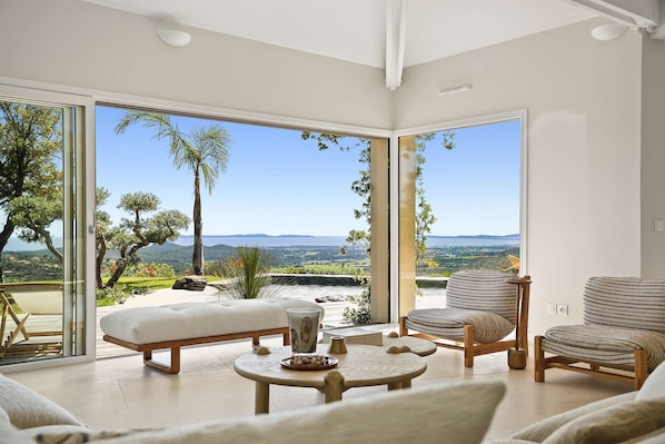 A modern living room with a large glass door leading to a scenic outdoor view, a cushioned bench, a round table, and striped armchairs. - Air-conditioned dream villa with lagoon-style pool and sea view for 6 guests in La Londe-les-Maures (LA LONDE LES MAURES)