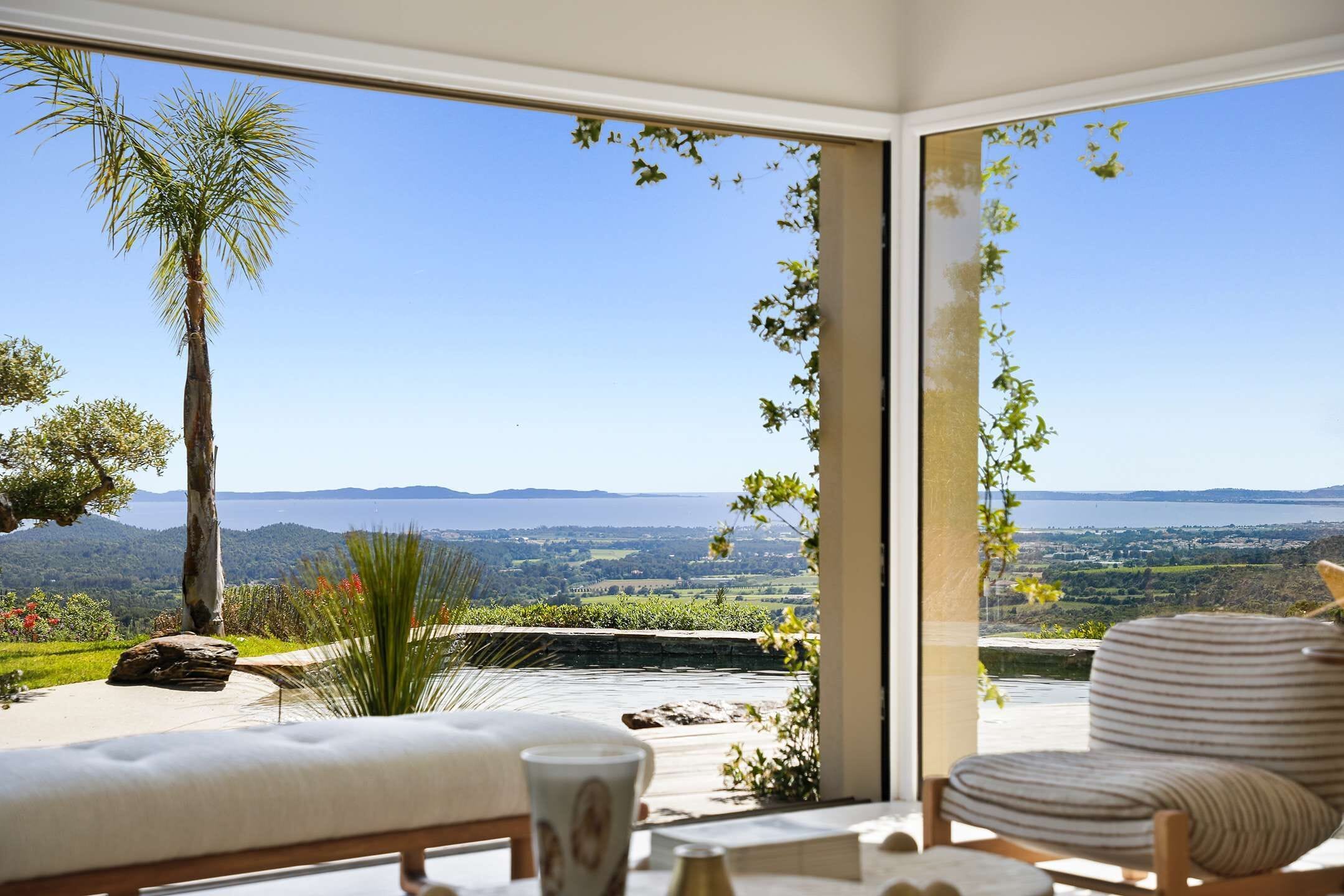 A room with a large window, a sofa, a chair, and a view of a landscape with hills and a body of water.