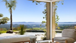 A room with a large window, a sofa, a chair, and a view of a landscape with hills and a body of water. - Air-conditioned dream villa with lagoon-style pool and sea view for 6 guests in La Londe-les-Maures (LA LONDE LES MAURES)