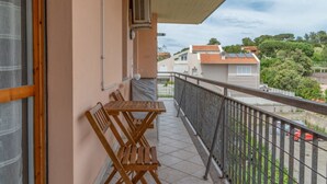 A balcony with a table and chairs, a window, and a view of neighboring buildings and greenery. - APPARTAMENTO SOTTOBOMBA (Portoferraio)