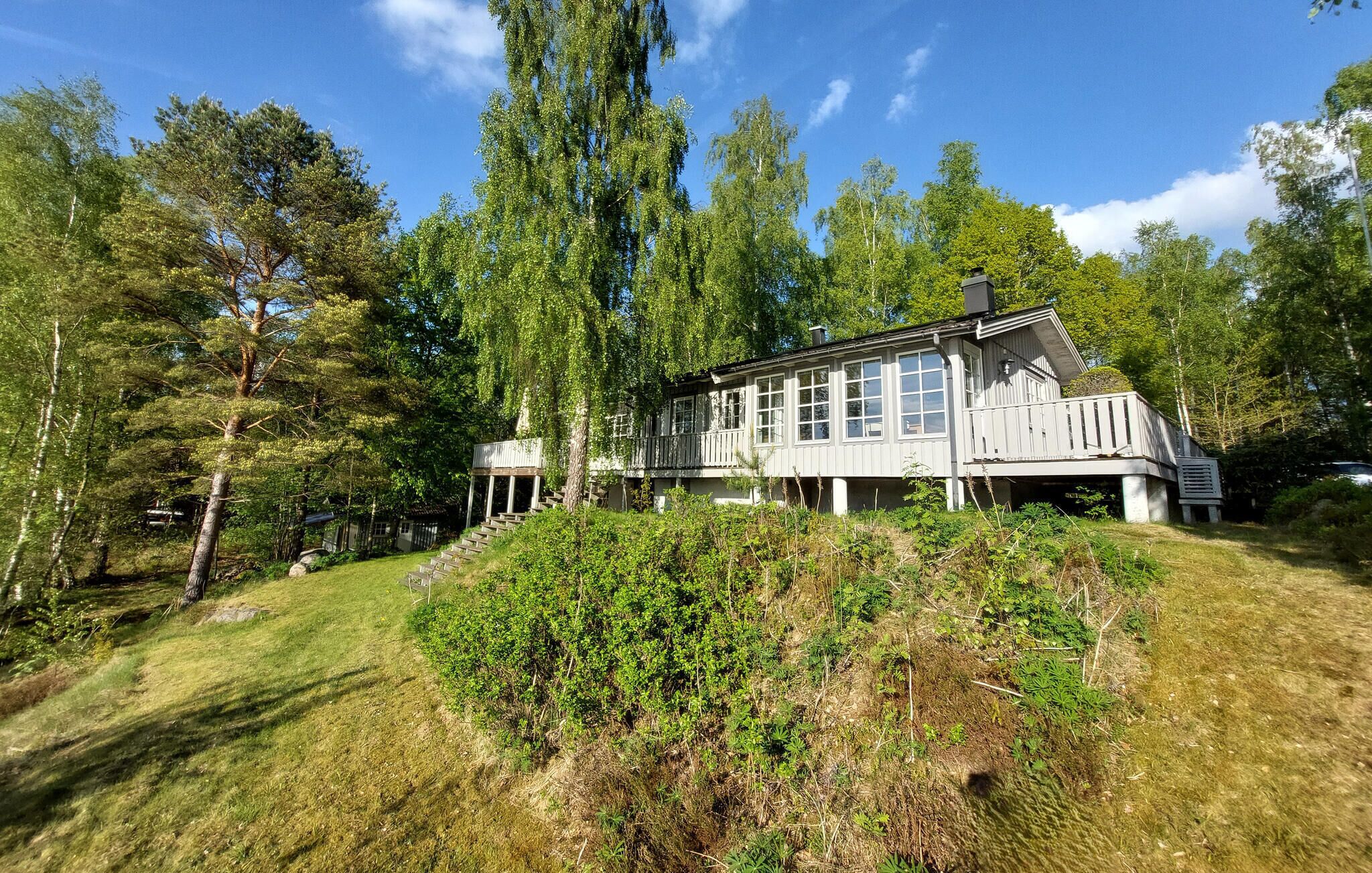 A large, single-story house with a covered porch and a small deck, surrounded by dense trees and a grassy area.