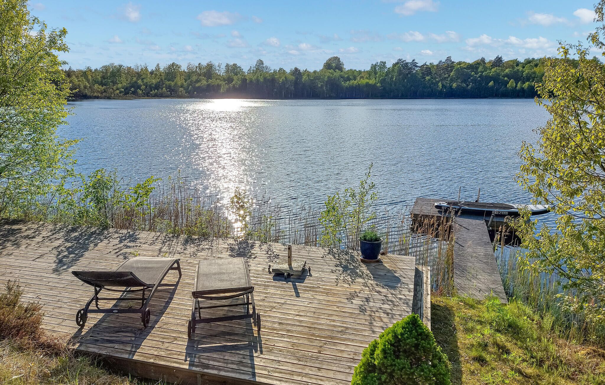A wooden deck with two sun loungers and a small table, overlooking a calm lake with a dock and trees in the background.