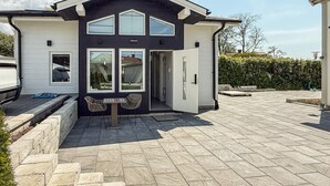 Uma casa moderna de dois andares com fachada escura, um alpendre coberto com mesa e cadeiras, e uma entrada de garagem que leva a um carro estacionado.