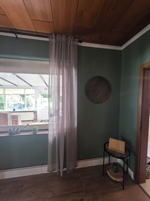 Una habitación con techo de madera, pared verde, un objeto decorativo redondo, una silla y una ventana con vistas al exterior.