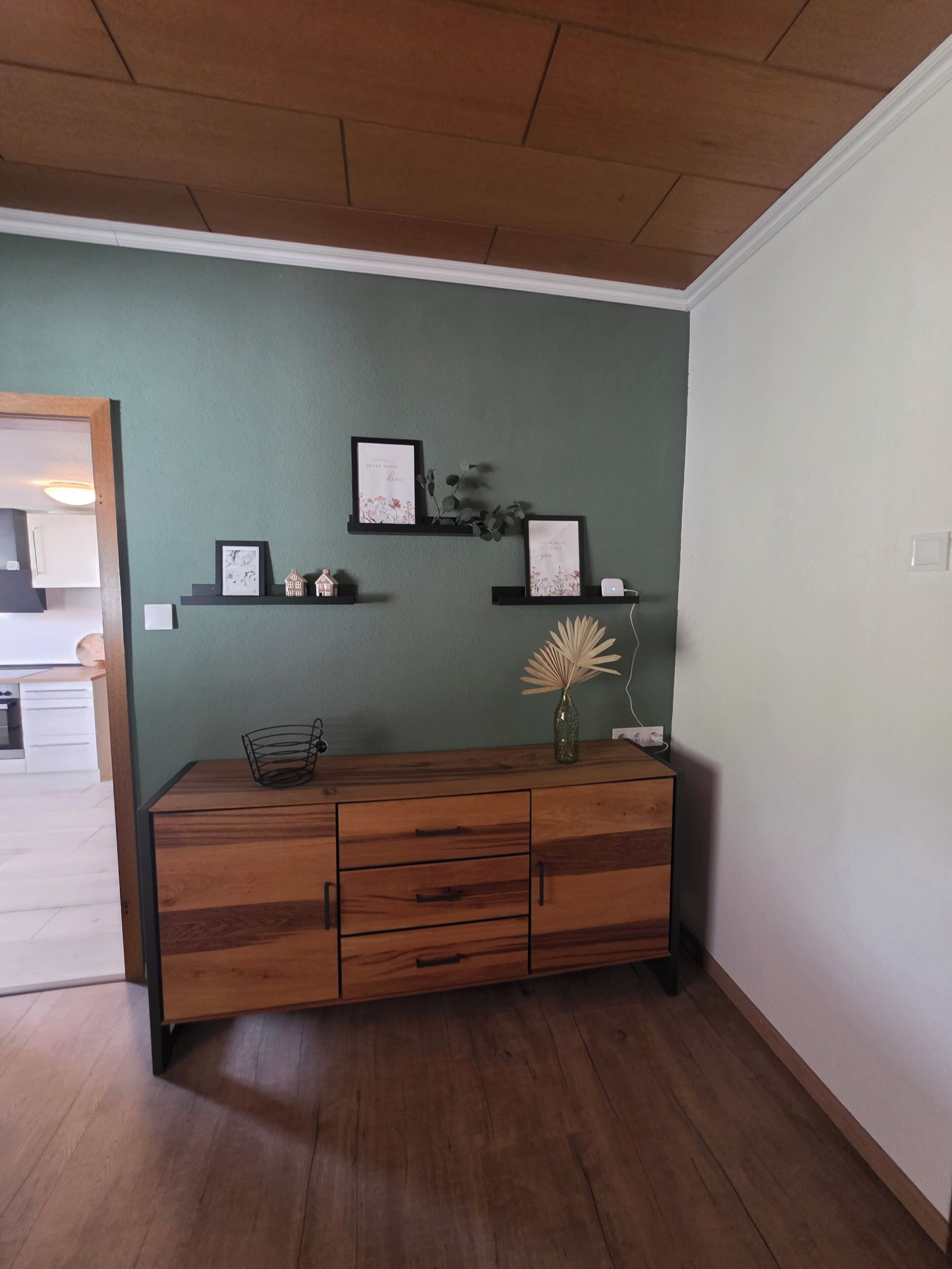 Een kamer met een houten kast, twee wandplanken met decoratieve objecten, en een vaas met gedroogde bloemen.