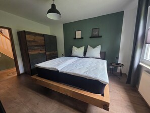 A bed with a wooden frame, a white mattress, and two pillows. There is a small table with a lamp beside the bed. The room has a green wall and a window with curtains.