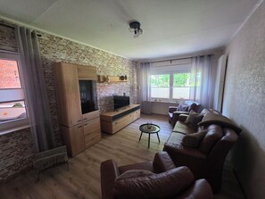 A living room with a brick wall, wooden furniture, a TV, a sofa, and a window with curtains.