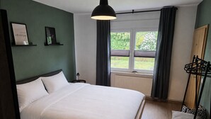 Ein Schlafzimmer mit einem Bett, einem Fenster mit Vorhängen, einem Heizkörper und einem an der Wand befestigten Regal mit gerahmten Bildern.