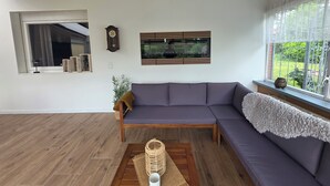 Ein modernes Wohnzimmer mit einer Eckcouch, Holzboden, Deckenbalken und einer Wanduhr. Ein Fenster bietet Blick ins Grüne.