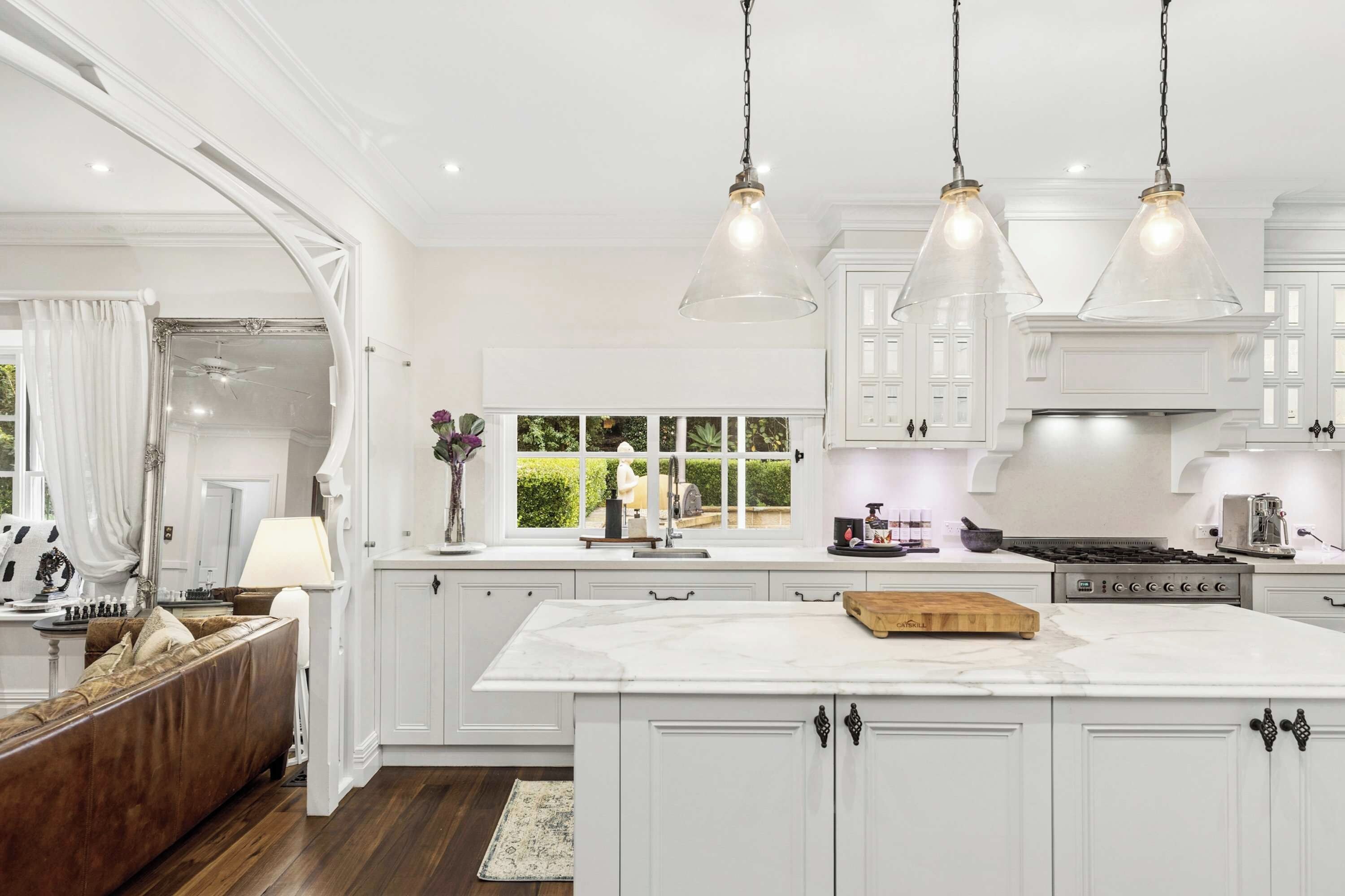 A spacious kitchen with white cabinetry, a large island, pendant lighting, and a view of greenery outside.