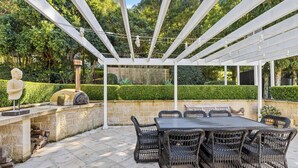 En udendørs terrasse med pergola, stenvæg og et bord med fletstole.
