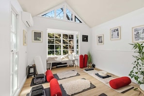 Ein Zimmer mit schrägen Wänden, großen Fenstern, Yogamatten, einem roten Kissen, einem kleinen Tisch und gerahmten Bildern an den Wänden.
