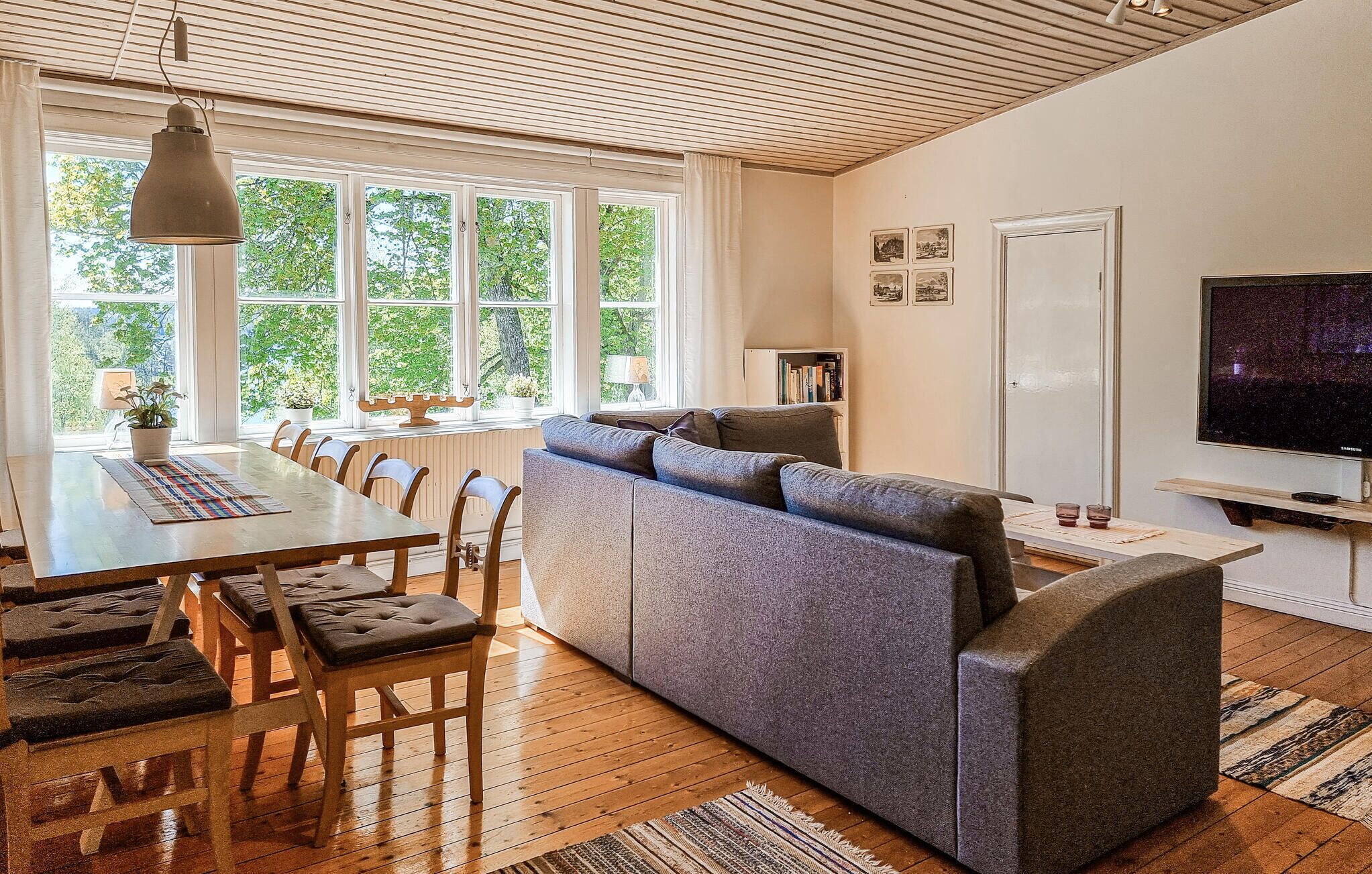 A modern living room with a wooden ceiling, a large window with curtains, a grey sofa, a dining table with chairs, and a flat-screen TV.