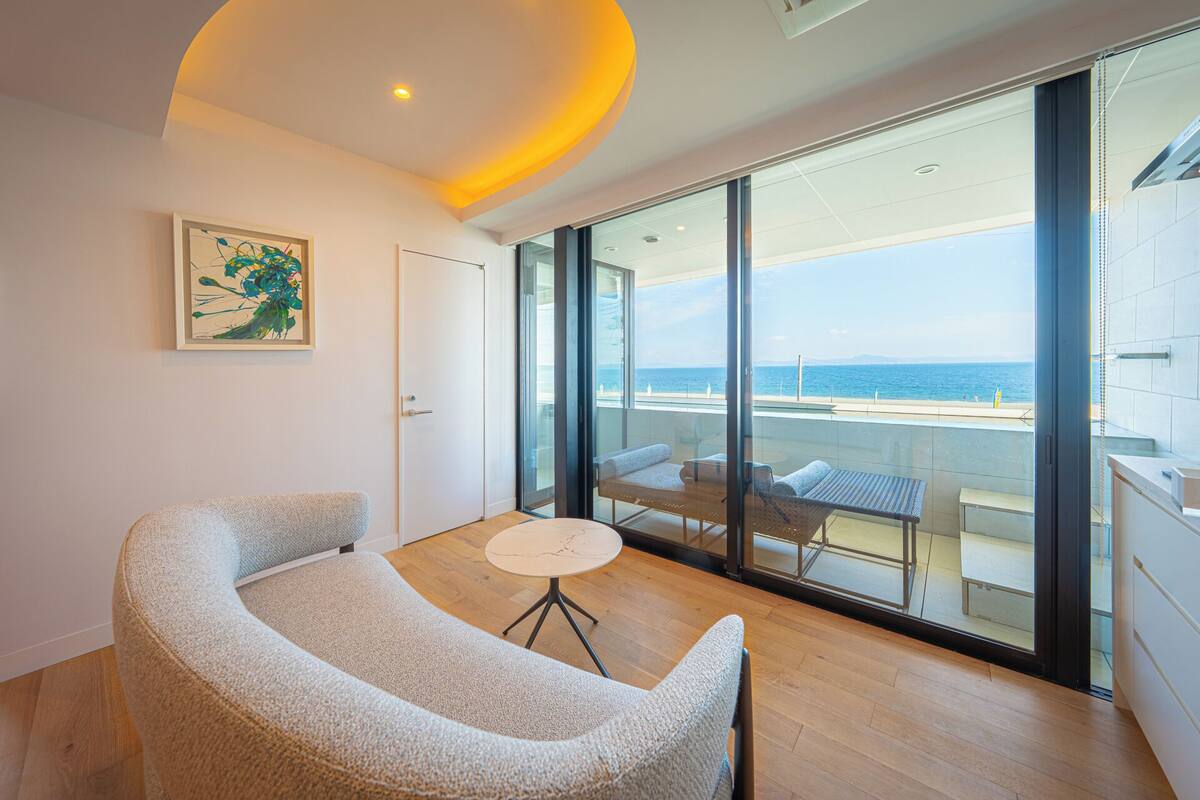 Una habitación moderna con un sofá curvo, una pequeña mesa redonda y un amplio ventanal corredero que da a un balcón con vistas al mar.
