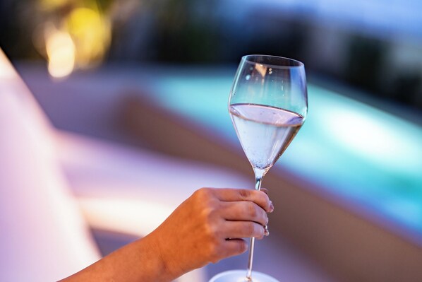 Una mano sostiene un vaso transparente con un líquido, posiblemente agua, y un fondo desenfocado que sugiere una piscina o entorno exterior.