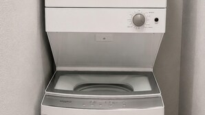A front-loading white washer and dryer in a corner with a wooden floor and light-colored walls. - Cozy 1BR Near Med Center, NRG & Downtown (Houston)