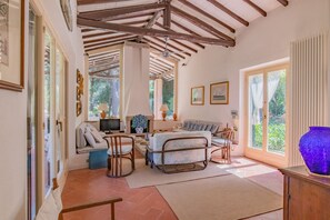 Ein geräumiges Wohnzimmer mit hoher Decke, einer Sofagarnitur, einem Fernseher und einem großen Fenster mit Blick ins Freie.