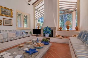 A living room with a sofa, a TV, a fireplace, and a window with a view of trees. - Villa Thalèa Fetovaia (Campo nell'Elba)