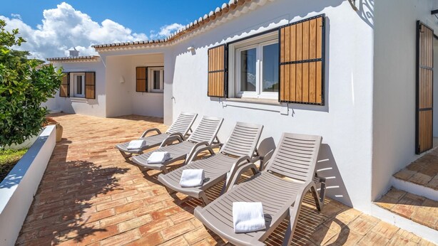 Ein weißes Gebäude mit einem Ziegeldach, Holzläden und einer Terrasse mit Liegestühlen.