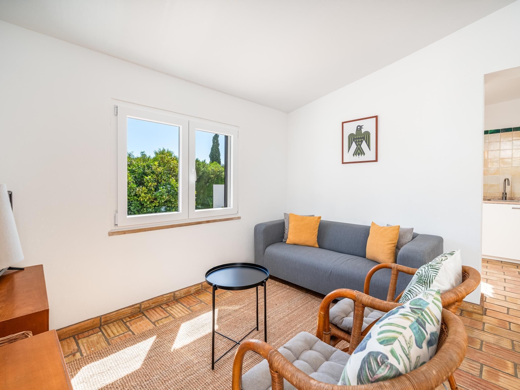 A living room with a grey sofa, two yellow cushions, a wicker chair, a small round table, and a framed picture on the wall.
