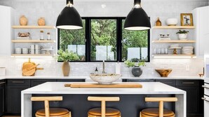 A modern kitchen with a central island, wooden stools, pendant lighting, and a view of greenery outside.