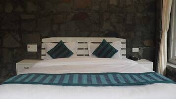 A bed with white bedding and two pillows, a stone wall, a nightstand with a lamp, and a window with curtains.