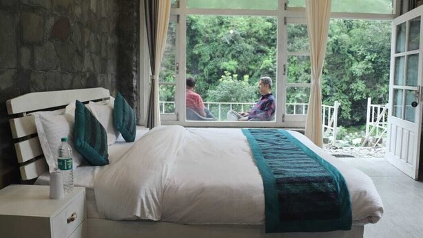 A bedroom with a large window, a bed, a nightstand, and a view of a balcony with people outside.