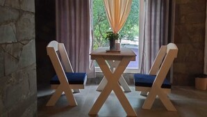 A room with a stone wall, a window with curtains, a table with a plant, and two chairs.