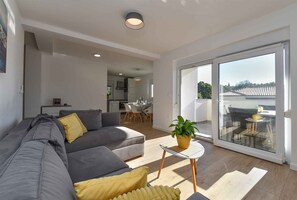 A modern living room with a grey sofa, a small white table with a plant, and a sliding glass door leading to an outdoor area. - Holiday flat with full amenities (Štinjan)