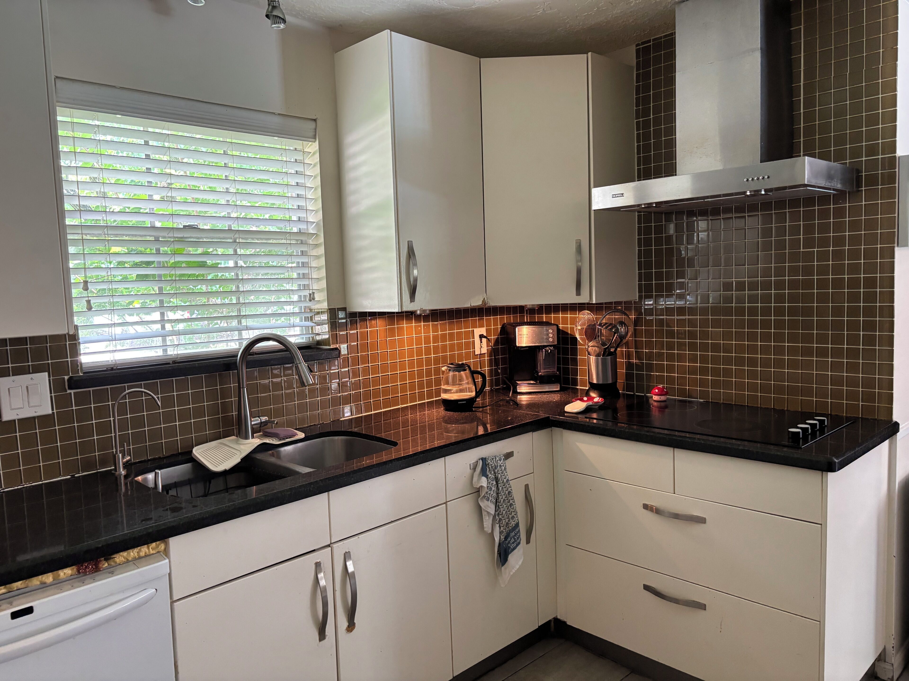 A modern kitchen with white cabinets, a black countertop, a stainless steel range hood, and a window with blinds.