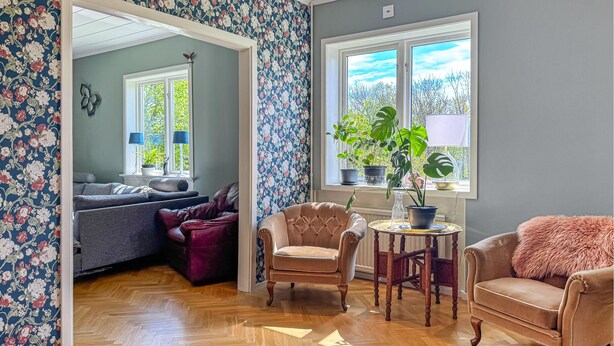 Ein Zimmer mit Fischgrätparkett, einer Couch, zwei Sesseln, einem kleinen runden Tisch und einem Fenster mit Blumentöpfen.