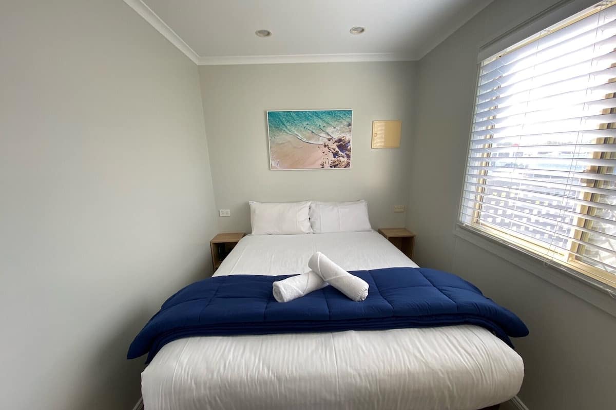 Una cama bien hecha con edredón azul y blanco, una mesita de noche, un interruptor de luz en la pared y una ventana con persianas.