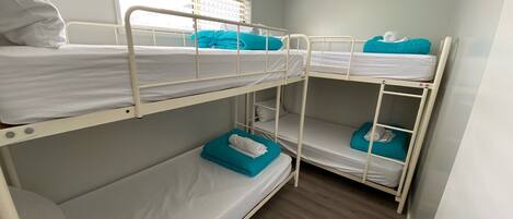 Habitación con literas, ropa de cama blanca y almohadas color turquesa, una ventana con persianas y suelo de madera.