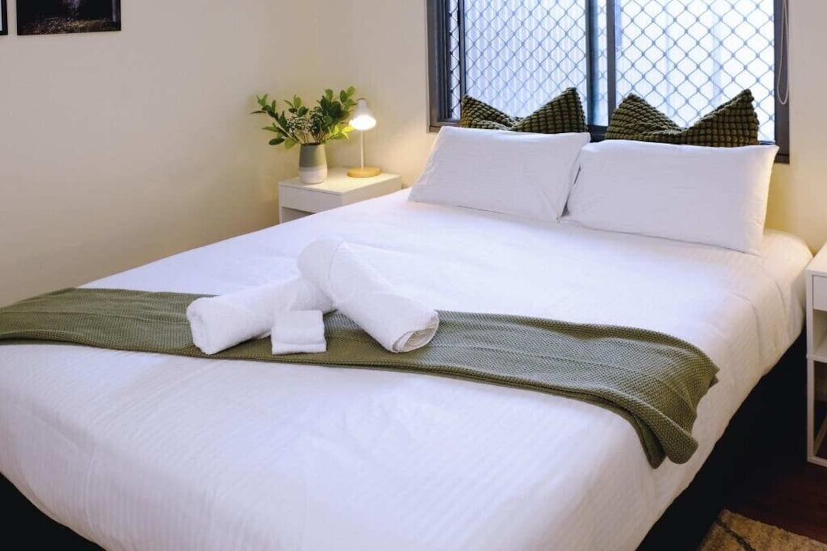 Una cama bien hecha con sábanas blancas, una manta verde y un par de toallas dobladas. Hay una mesita de noche con una lámpara y una planta en maceta.