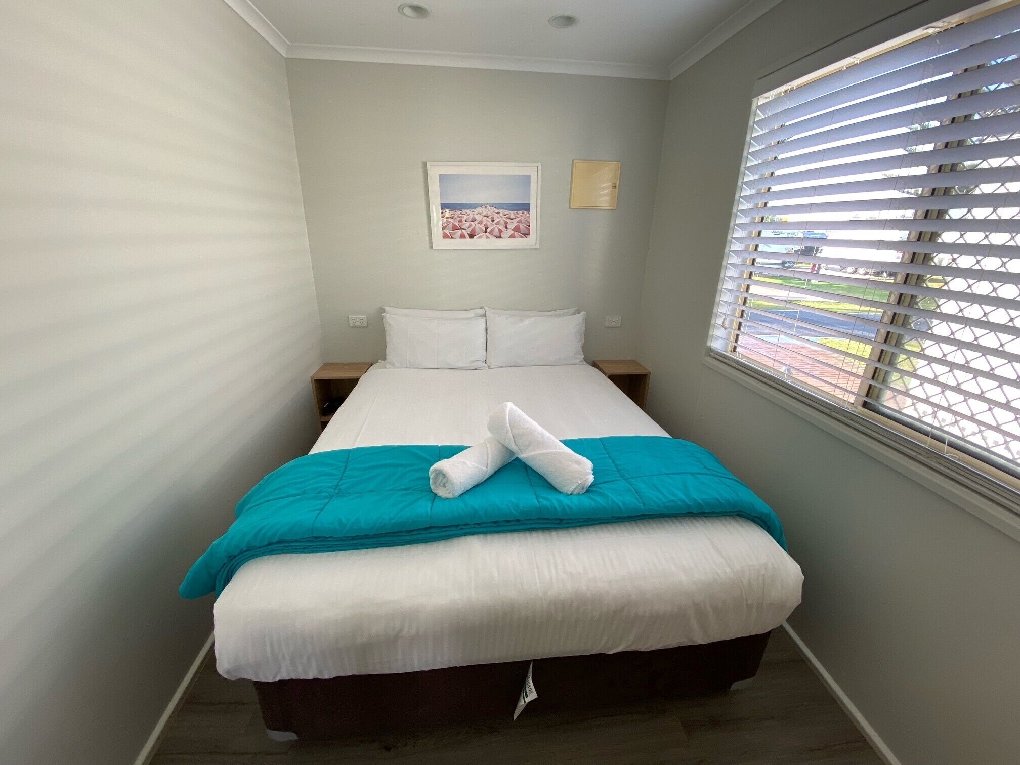 A single bed with a teal blanket, white pillows, and a rolled towel. There is a framed picture on the wall, a small wooden nightstand, and a window with blinds.