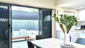 Un área de comedor con una mesa, sillas, un jarrón blanco con flores, una puerta corrediza de vidrio y una cocina al fondo.