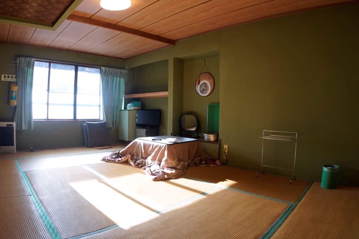 Ein Zimmer mit Tatami-Matten, einem Futon, einem Schreibtisch, einem Stuhl, einem Fernseher und einem Fenster mit Vorhängen.