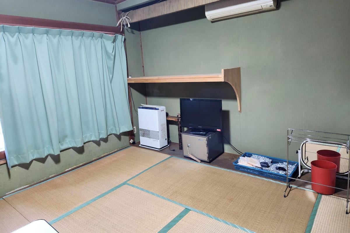 Ein Zimmer mit Tatami-Matten, einem Fernseher, einem kleinen Kühlschrank und einem Ventilator auf einem Regal.